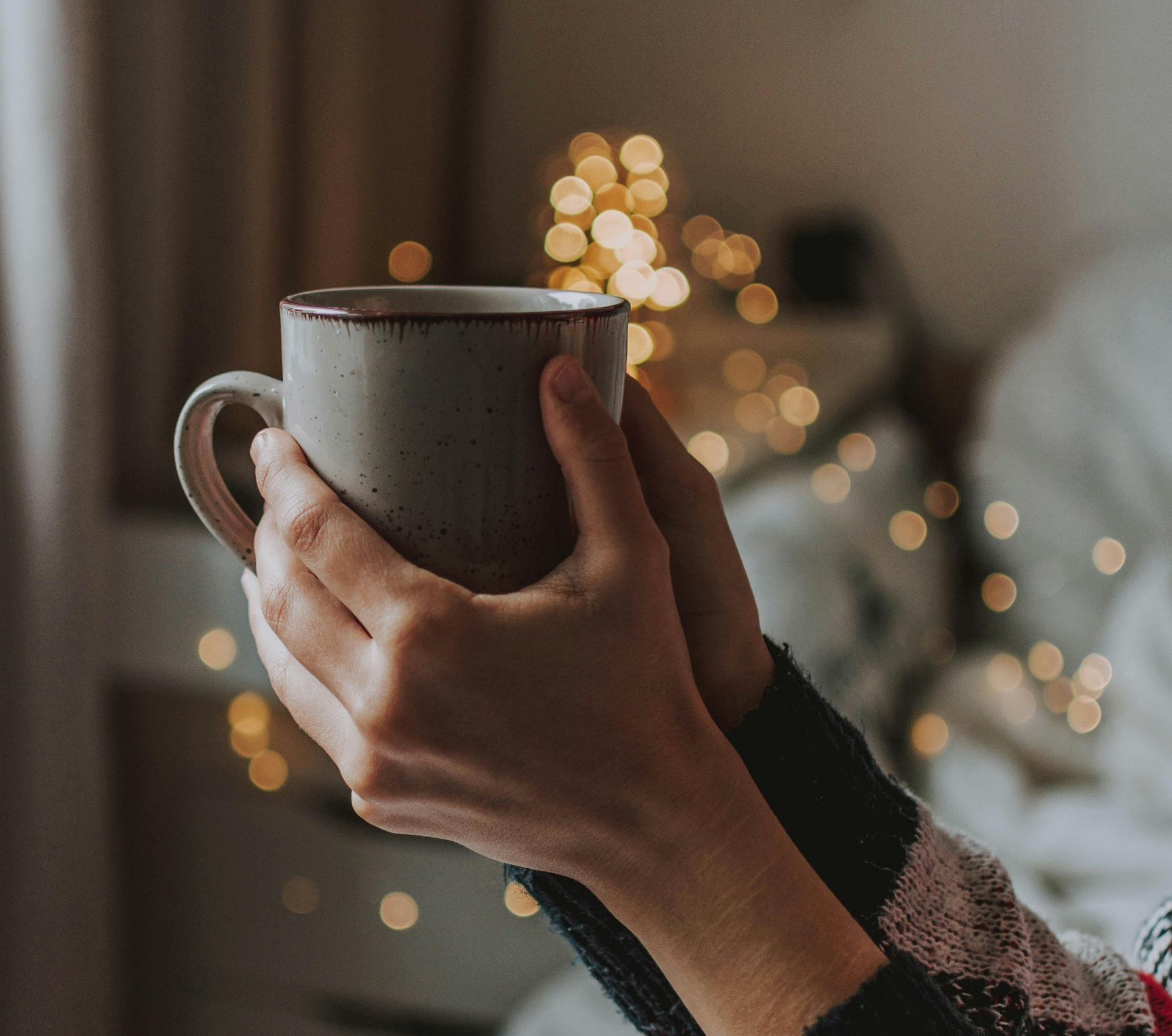Hands holding a warm mug, with fairy lights in the background.