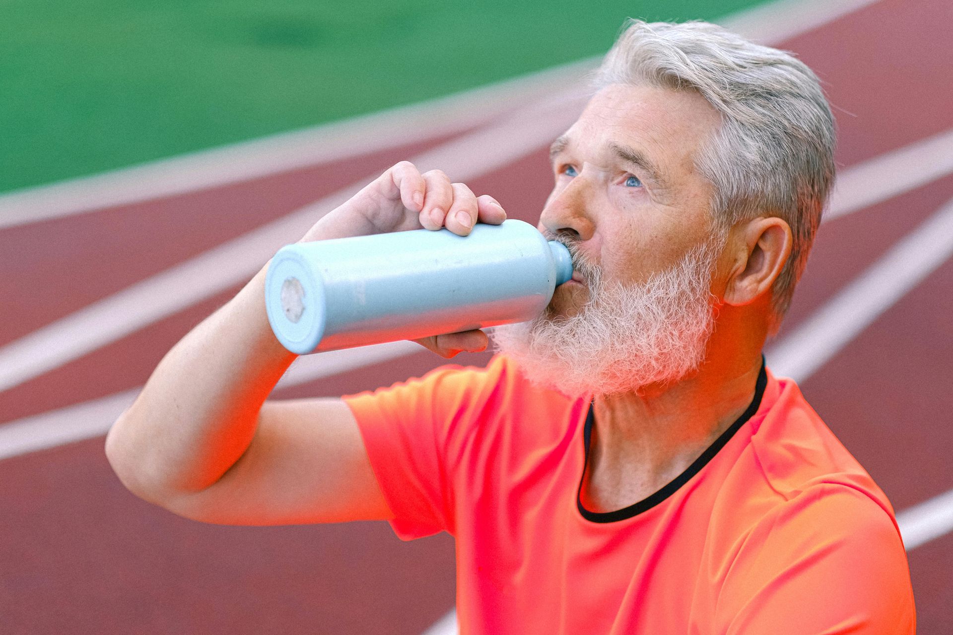 Senior man in orange shirt drinks from blue water bottle on running track.