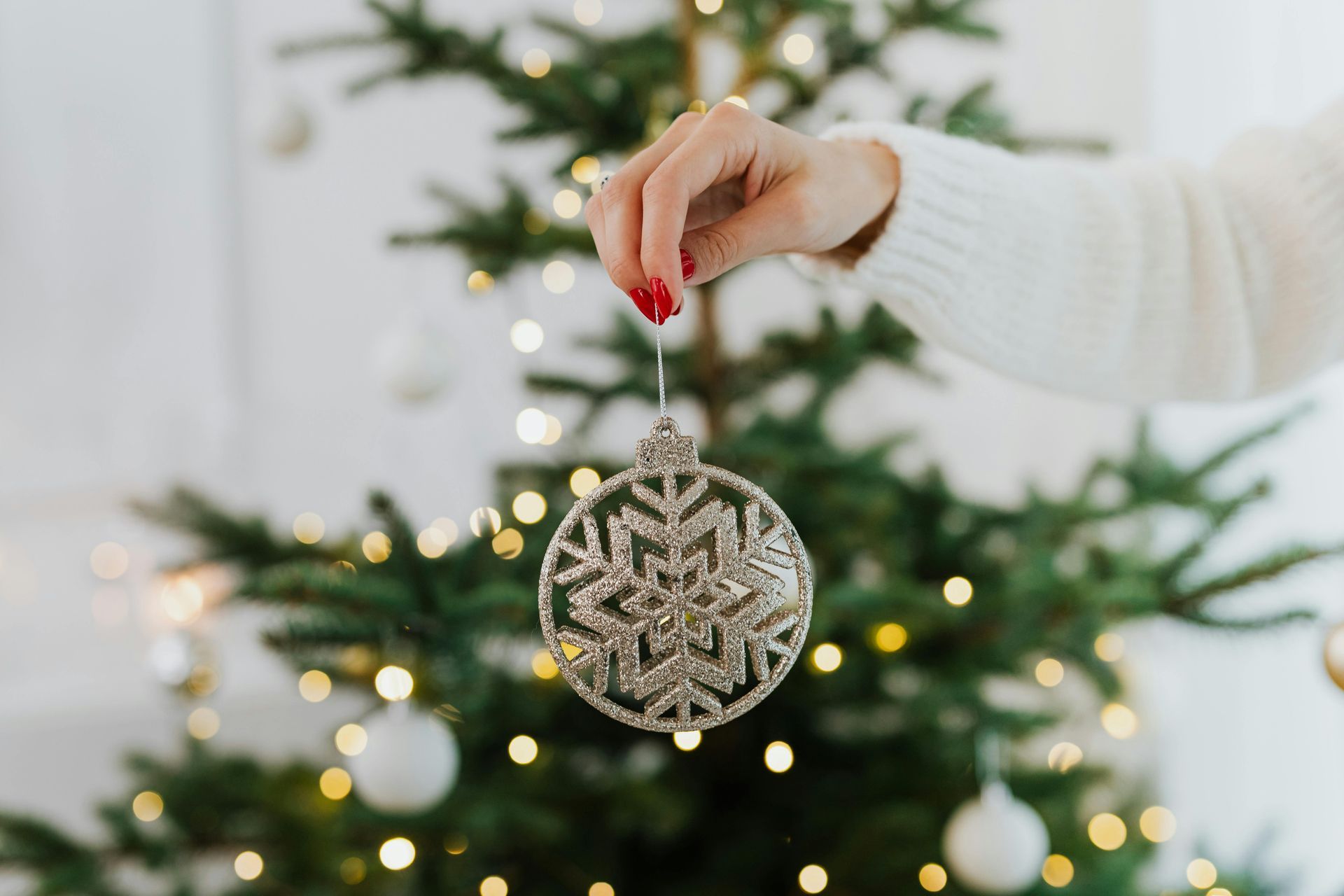 Hand holding silver snowflake ornament in front of a Christmas tree with white and gold lights.