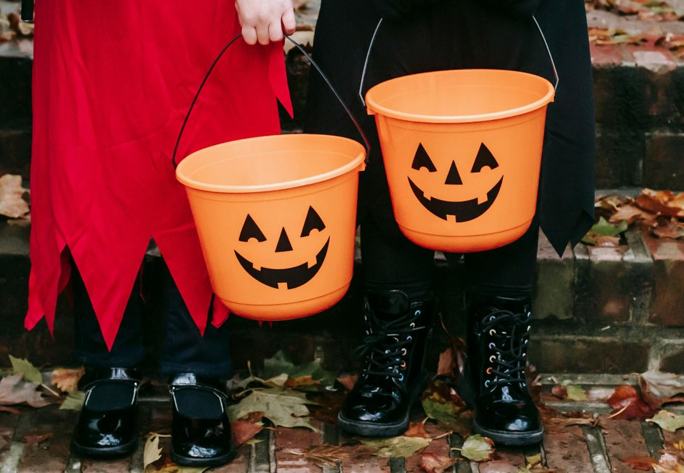 Two people in Halloween costumes holding pumpkin-shaped candy buckets on a brick walkway.
