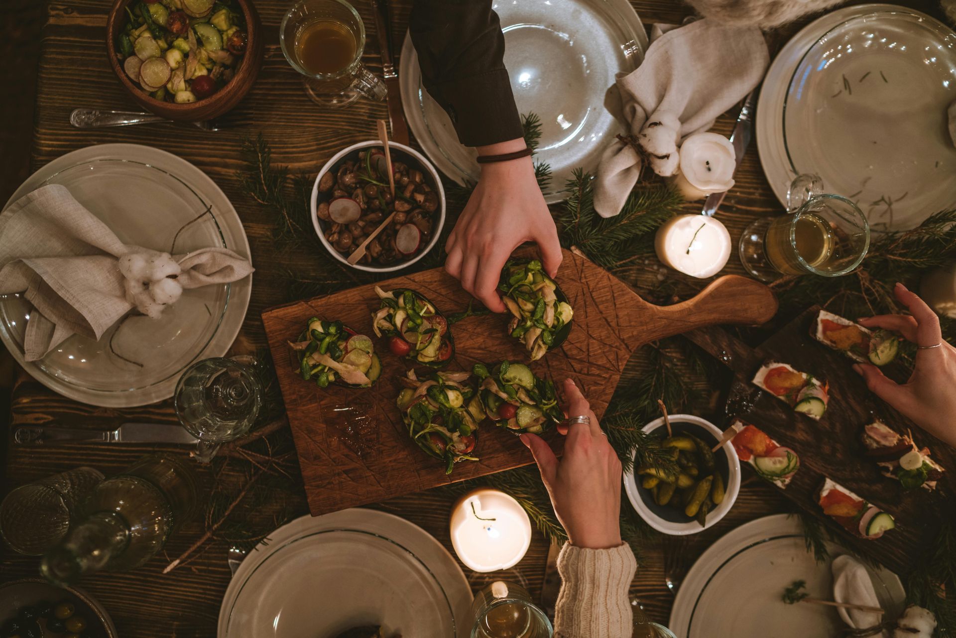 Overhead view of hands reaching for food on a wooden board at a dinner table set with plates, candles, and greenery.