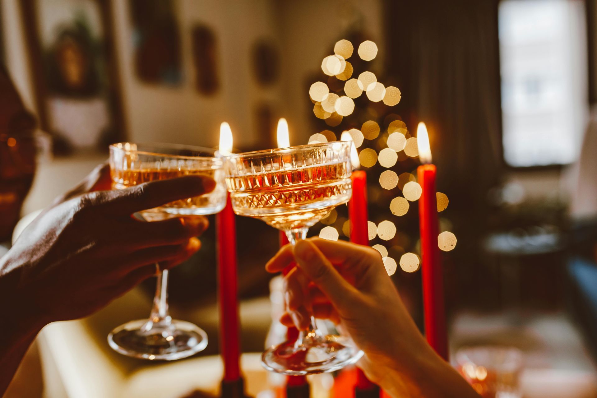 Hands clinking champagne glasses in front of lit red candles and a blurred Christmas tree.
