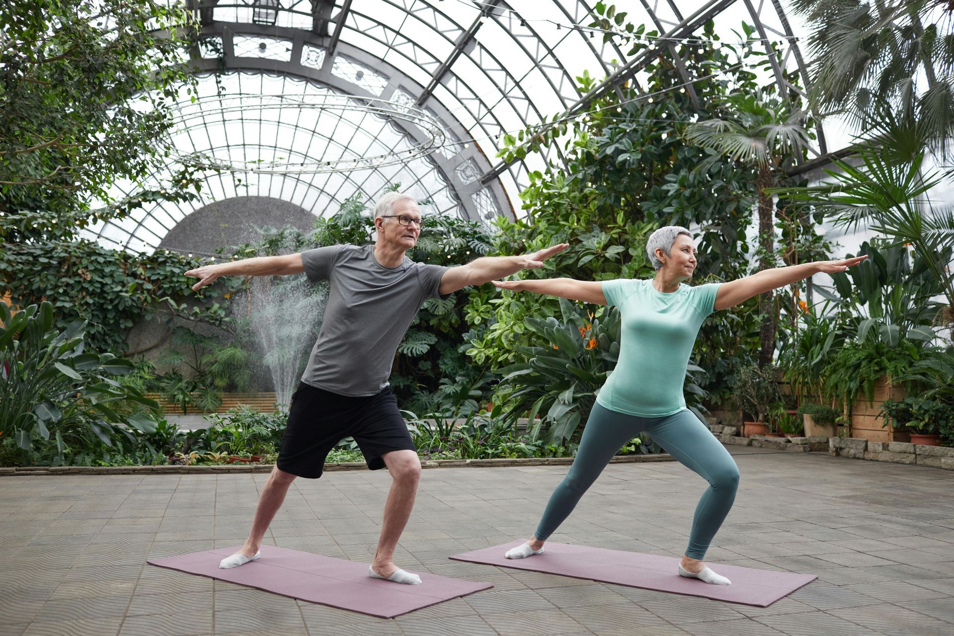 A man and woman do yoga outdoors on mats, extending arms, in a greenhouse-like setting.