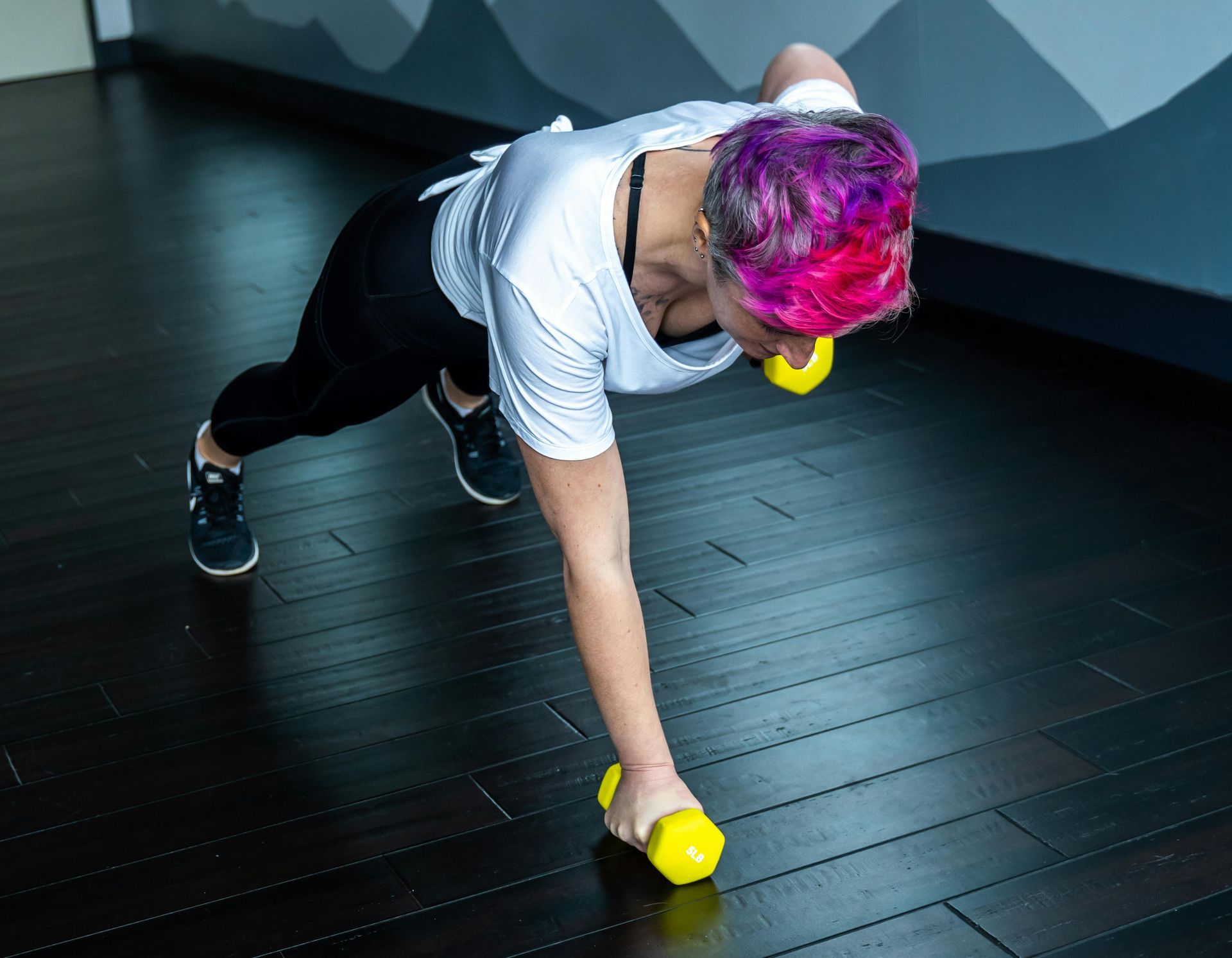 Woman in push-up position, lifting a yellow dumbbell. Wearing black pants and white shirt, purple hair, gym setting.