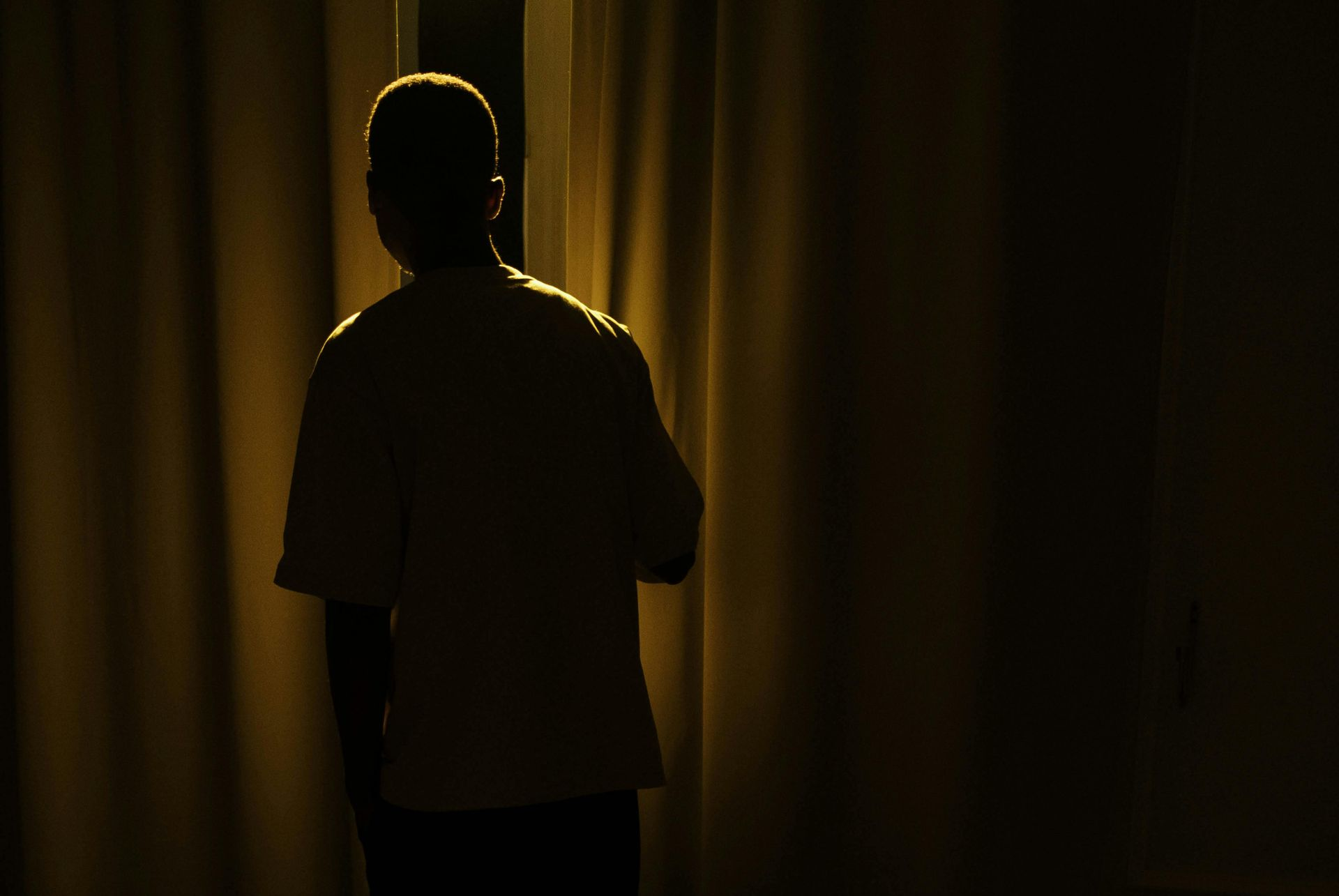 Man standing by curtains, in dark room; illuminated bed and curtains in gold light.