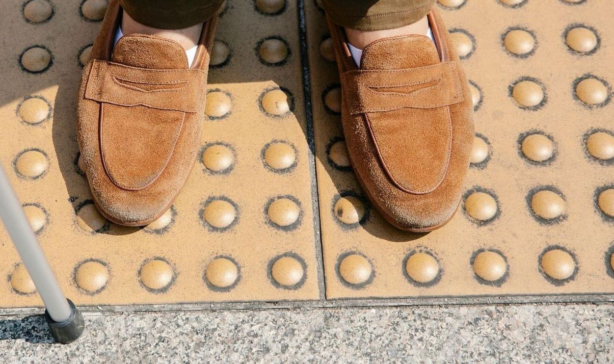 A person's feet wearing brown loafers on yellow tactile paving, with a white cane in the lower left.