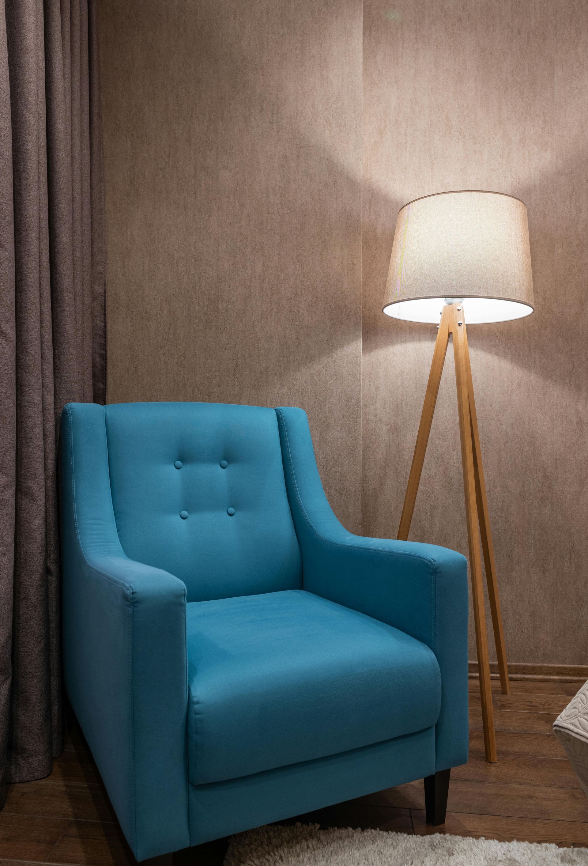 A teal armchair sits next to a wooden tripod floor lamp. The setting is a cozy room with brown walls.