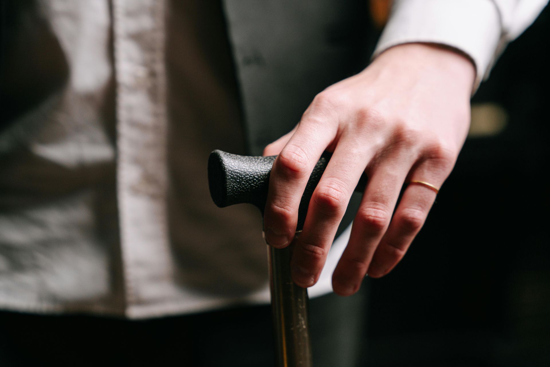 Man's hand resting on a cane; he wears a ring, dress shirt, and vest.
