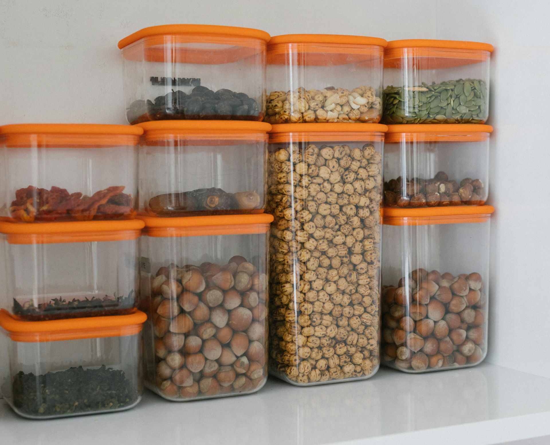 Clear food storage containers with orange lids filled with various dry goods, arranged on a white shelf.