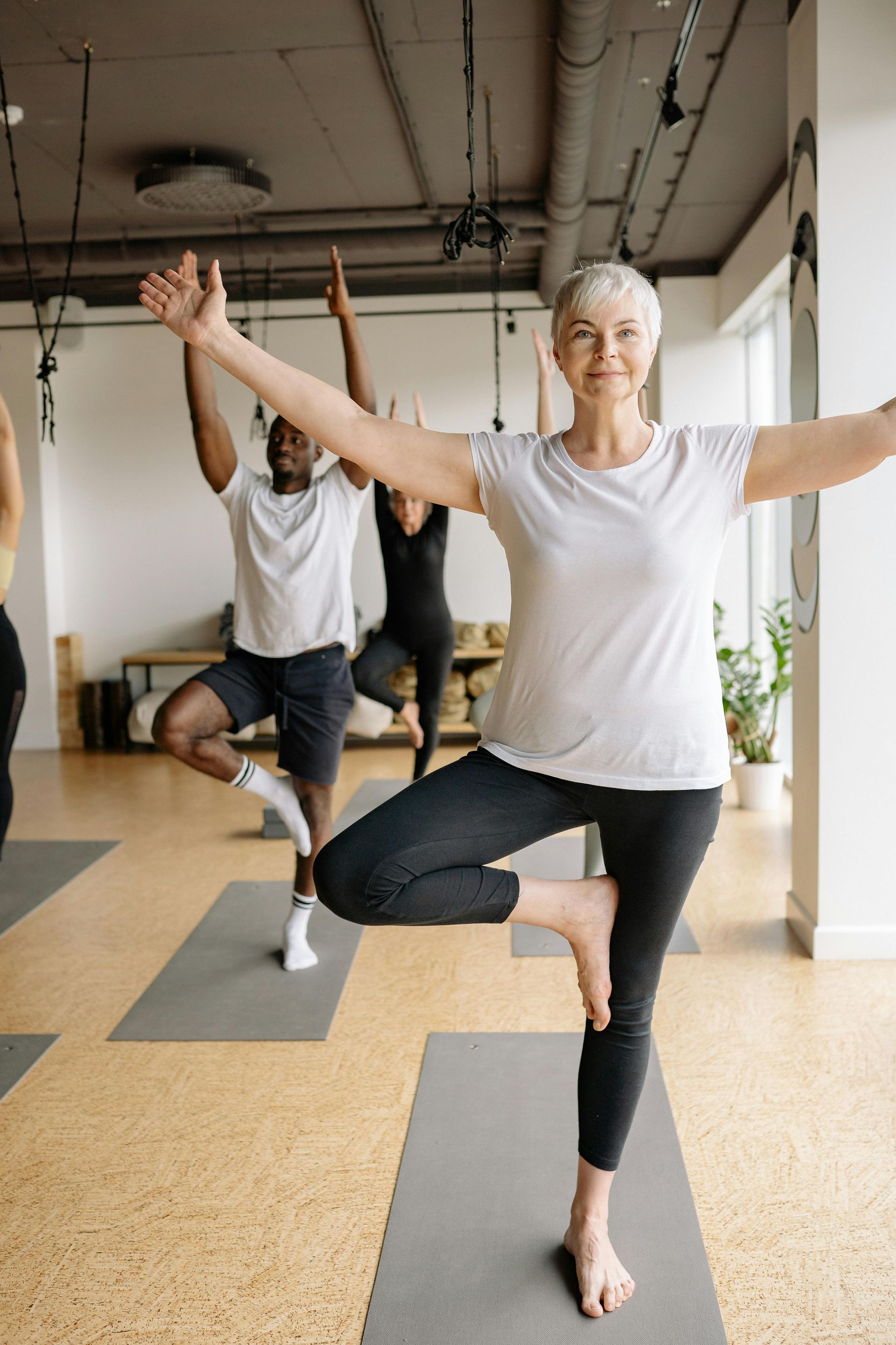 A woman with gray hair balances in a yoga pose with arms outstretched, in a studio with others practicing.