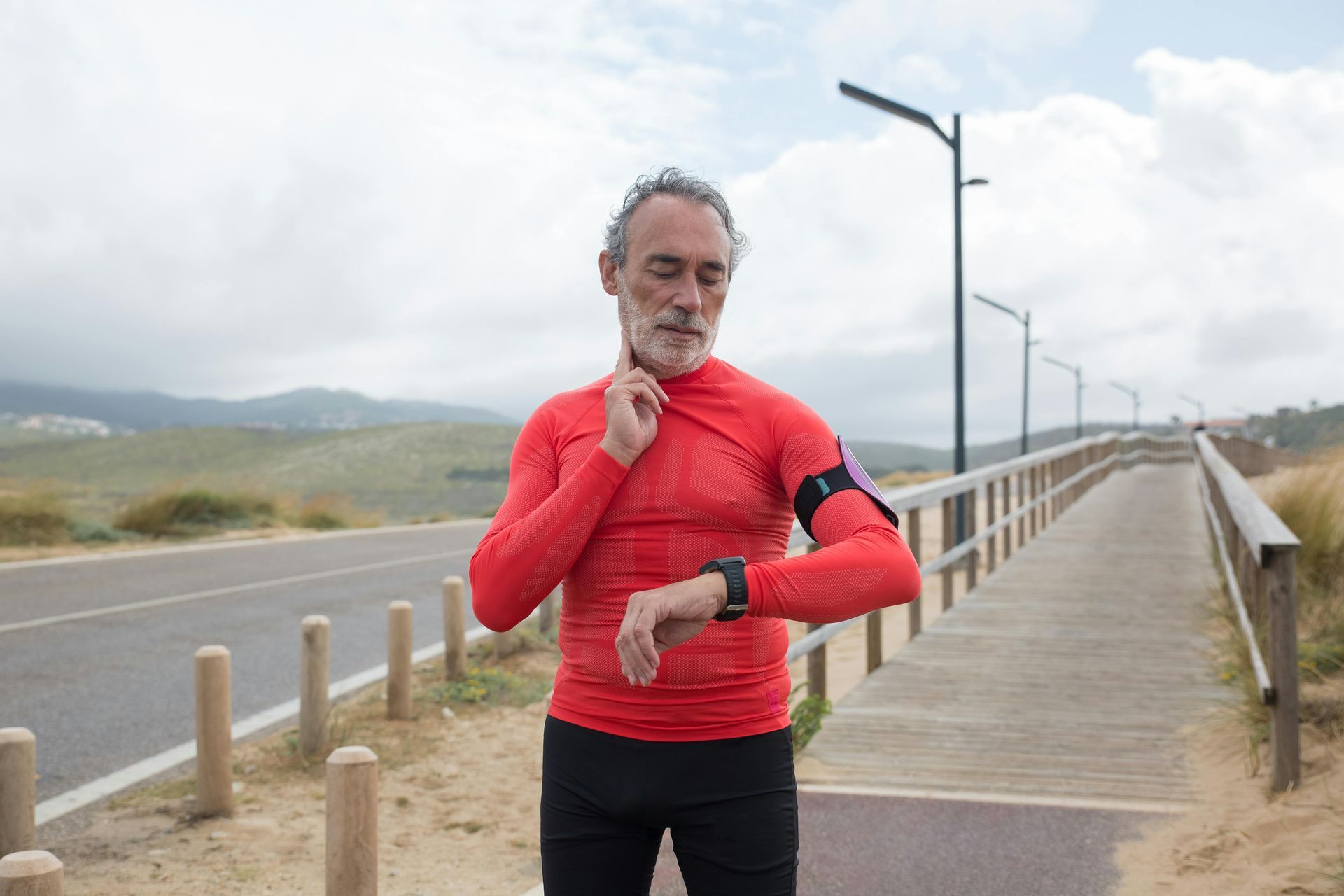 Older man in red shirt checking his pulse and smartwatch while standing on a boardwalk near a road.