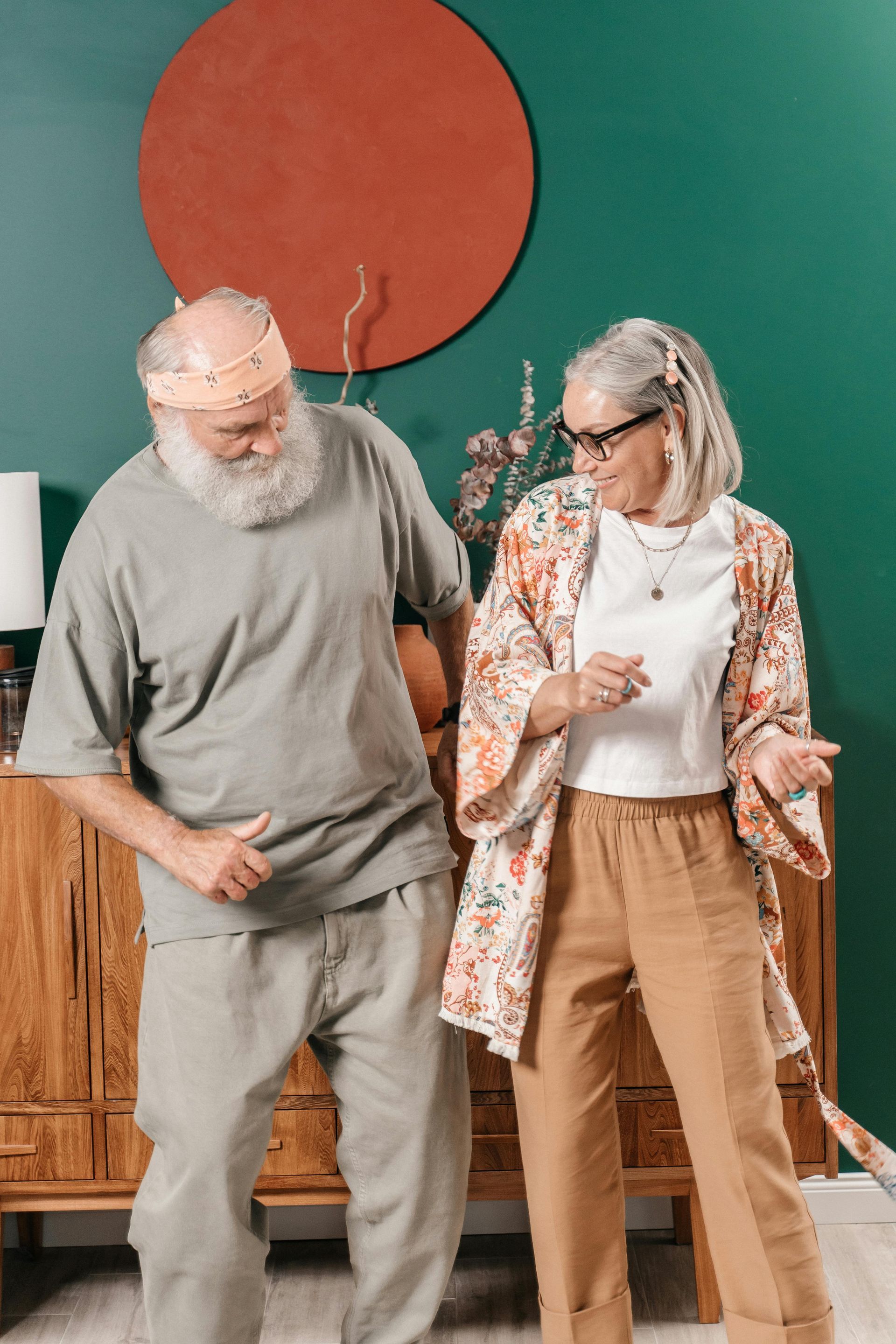 Two older adults dancing indoors in front of a green wall and a wooden cabinet.