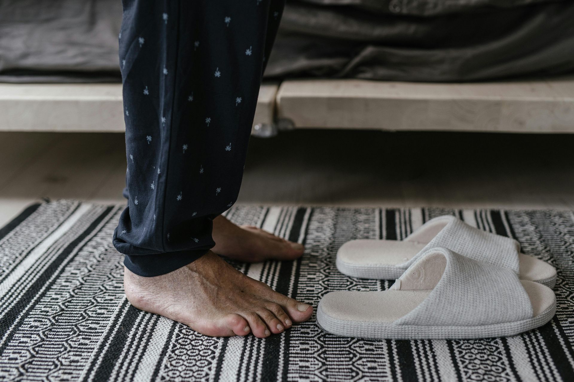 Person's bare feet next to white slippers on a striped rug. They wear blue pajama pants near a bed.