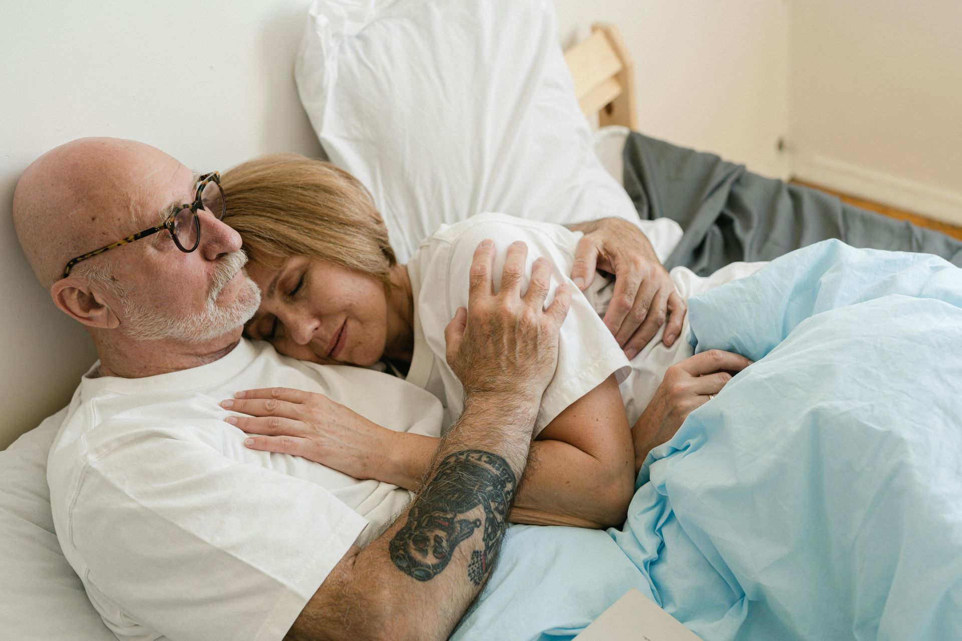 An older couple embraces in bed, the woman asleep on the man's chest. He wears glasses and has a tattoo on his arm.