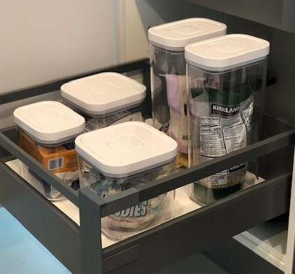 A drawer with organized pantry containers, white lids, various sizes, in a gray cabinet.