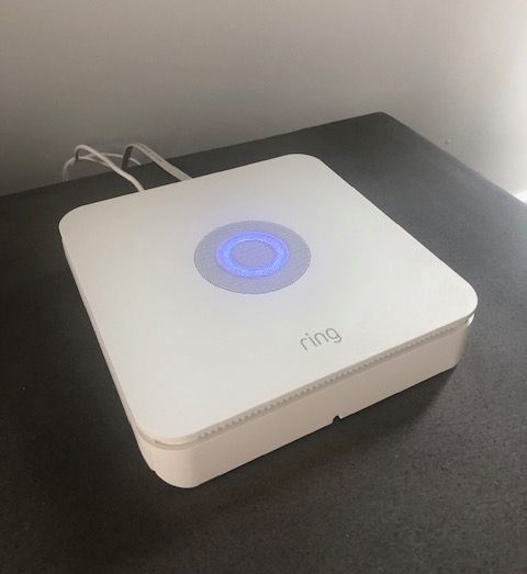 White Ring security system base station with blue glowing ring light, on a dark surface.