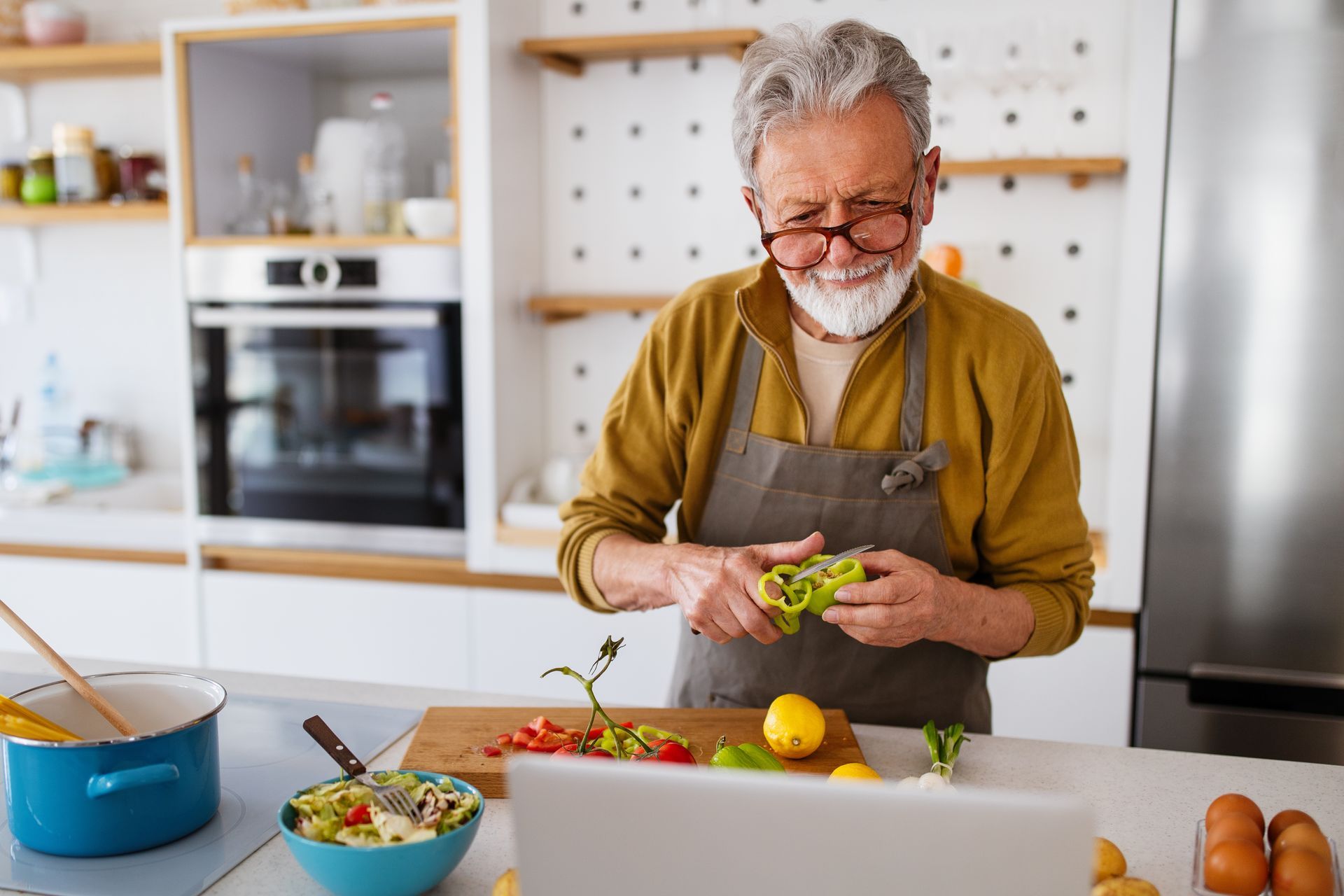 Elderly man in glasses and apron prepping vegetables in his kitchen while looking at a laptop.