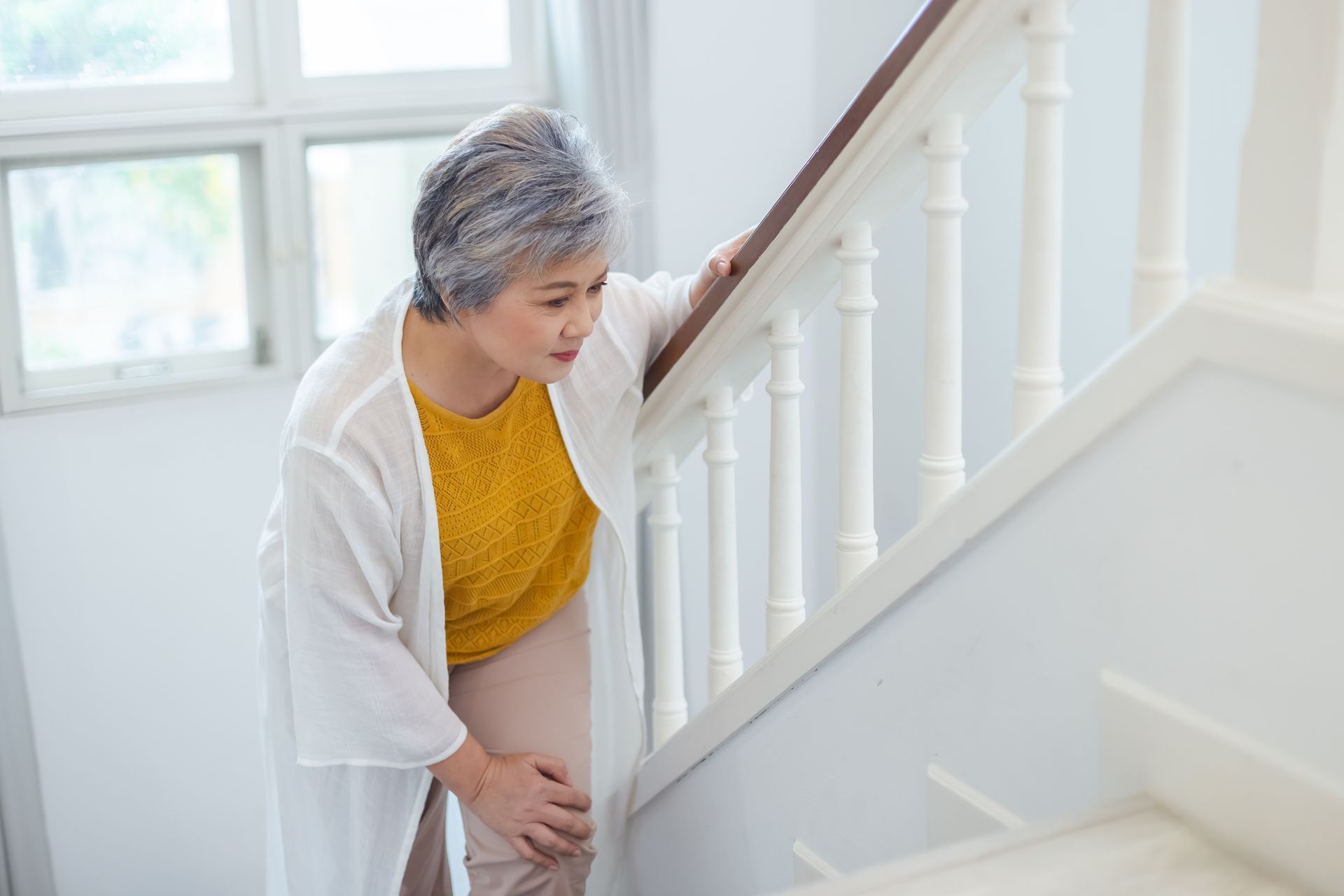 An older Asian woman struggles to climb stairs, clutching the handrail and holding her knee in pain.