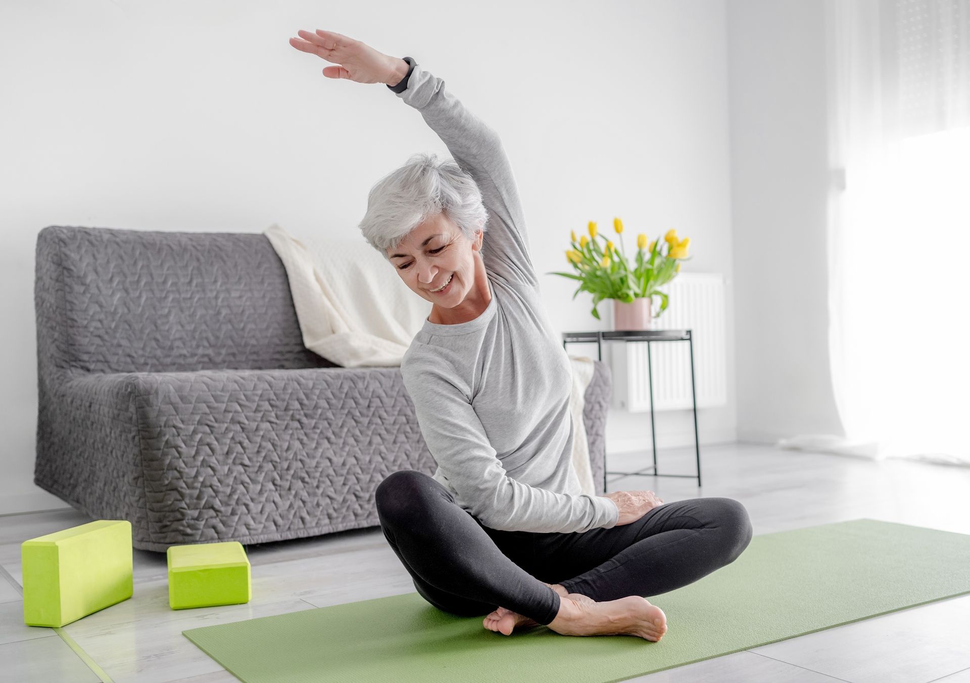 A smiling older woman in athletic wear stretches on a yoga mat indoors, reaching overhead.