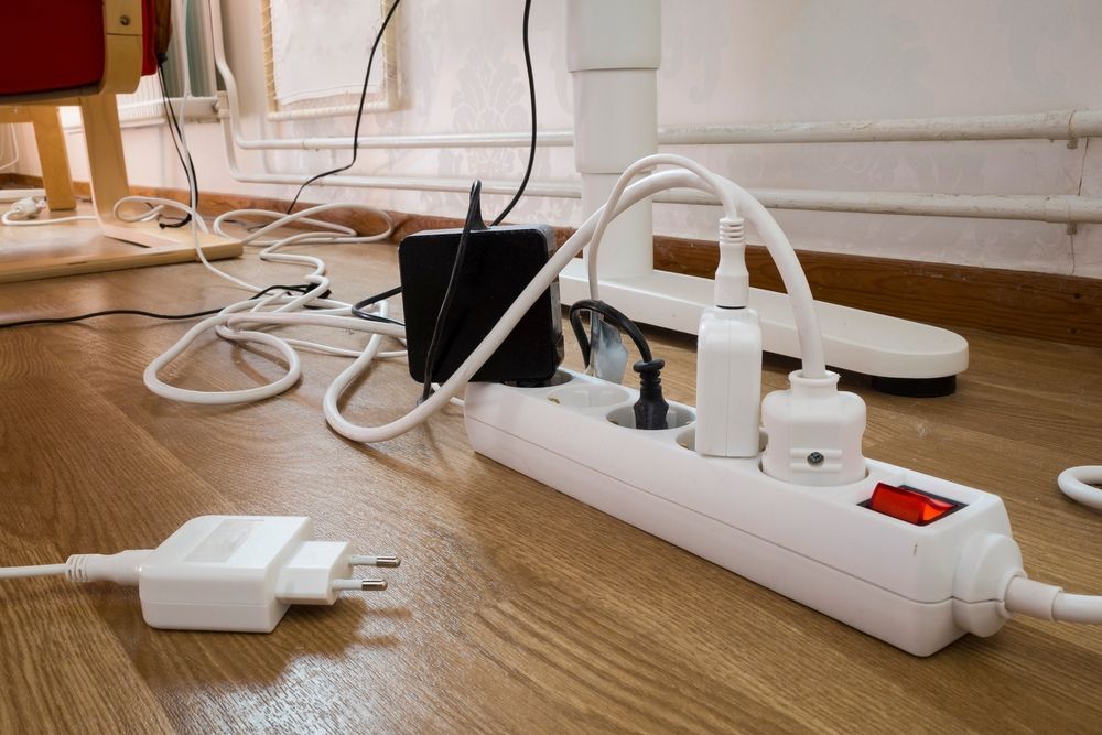 A white power strip on a wood floor is plugged into several devices. Cables and cords are sprawled around the strip.