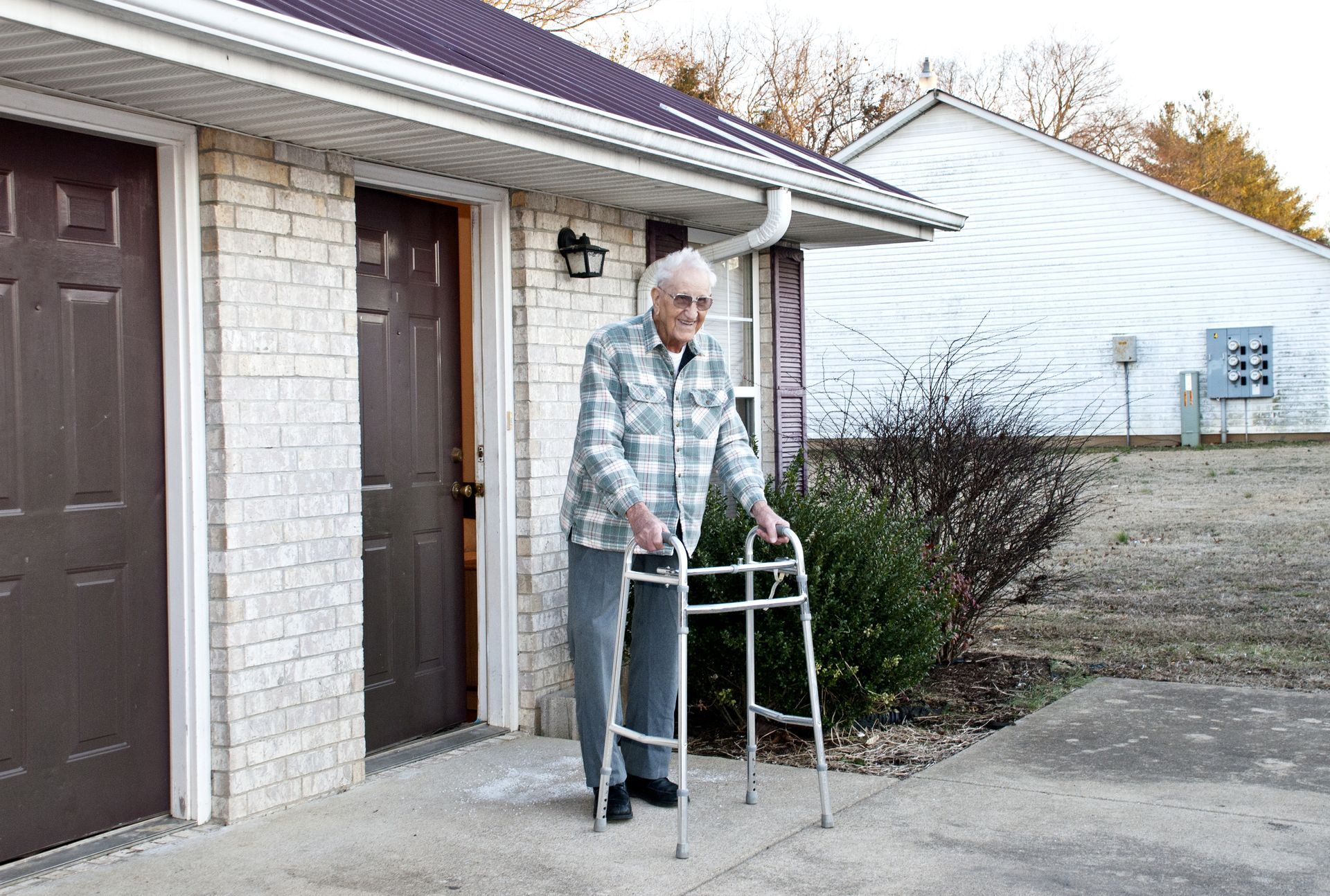 An elderly man with a walker stands outside a brick building with brown doors. He wears a plaid shirt and gray pants.