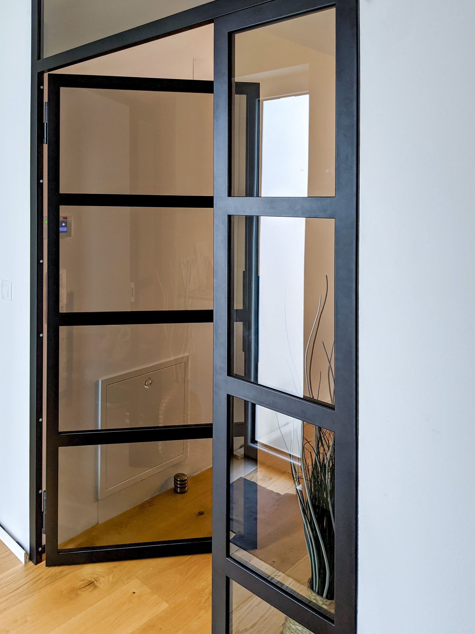 Offene, schwarzgerahmte Glastür in einem hellen Flur, dahinter sind eine weiße Wand und ein Heizkörper zu sehen.