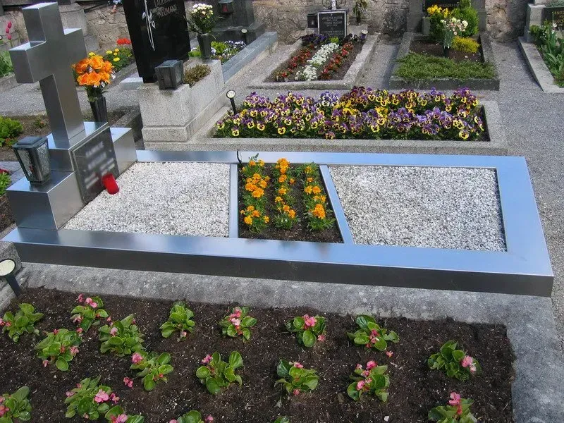 Grab mit weißem Kies und Blumenbeeten, eingefasst von grauem Stein auf einem Friedhof