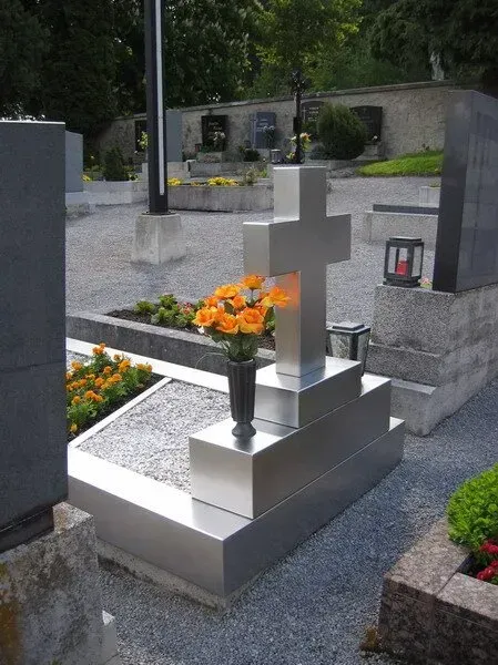 Silberner, kreuzförmiger Grabstein mit orangefarbenen Blumen auf einem Kiesfriedhof