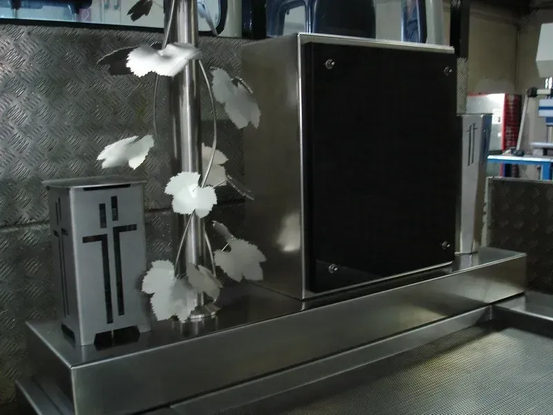 Metallene Gedenkurne mit weißen Blumen und silbernem Kreuz auf einem dunklen, reflektierenden Präsentationsständer