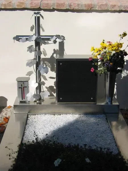 Gedenkstätte im Freien mit Blumen, Kreuz und kleinem Schrein auf einer Betonplattform im Sonnenlicht