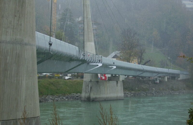 Hängebrücke im Nebel über einem Fluss, von der Seite betrachtet, mit Betontürmen und -kabeln.