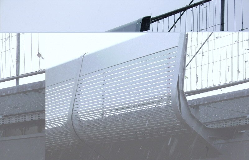Gebogene Fußgängerbrücke aus Metall mit perforierten Paneelen vor der Kulisse einer Stadtbrücke