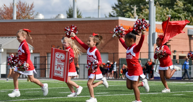 Cheerleader in red uniform performs a backbend on a green and red football field.