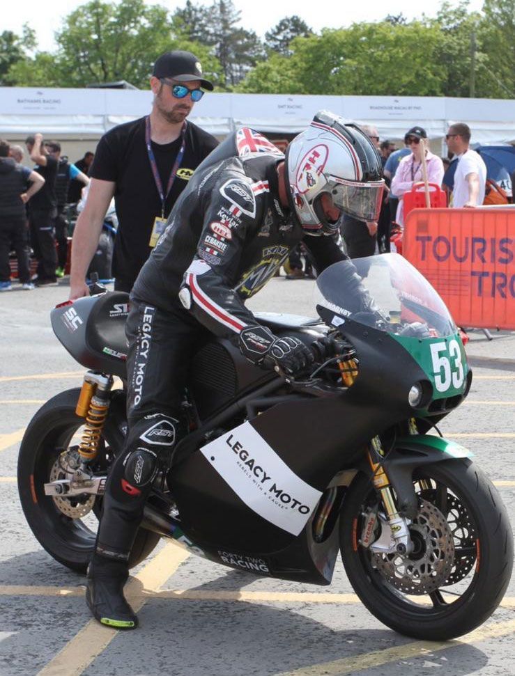Motorcycle racer on a black bike at the starting line, with crew and spectators in the background.