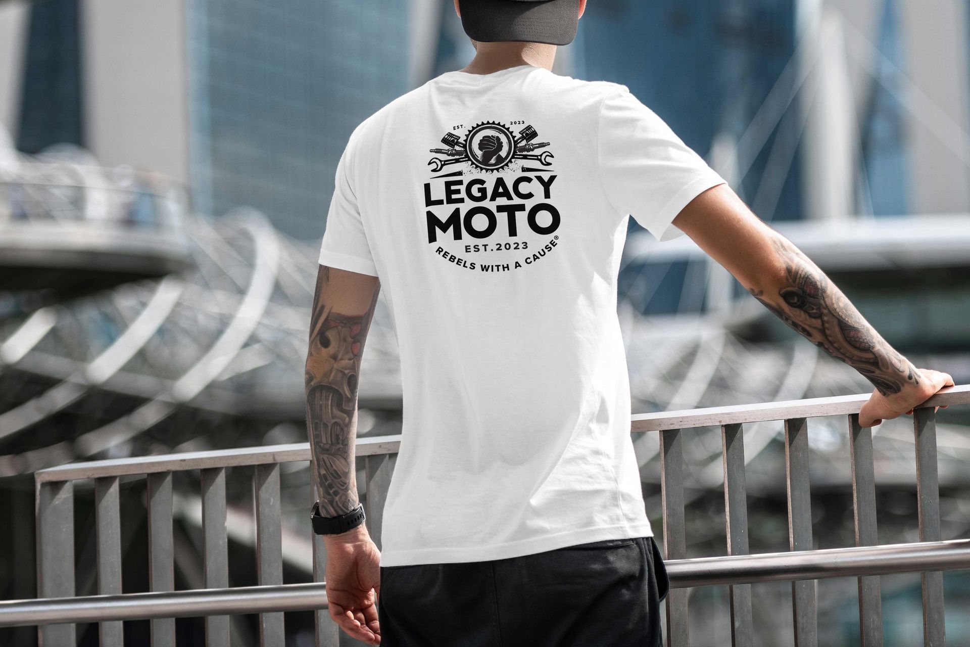 Tattooed person in a white T-shirt standing before a blurred stadium backdrop