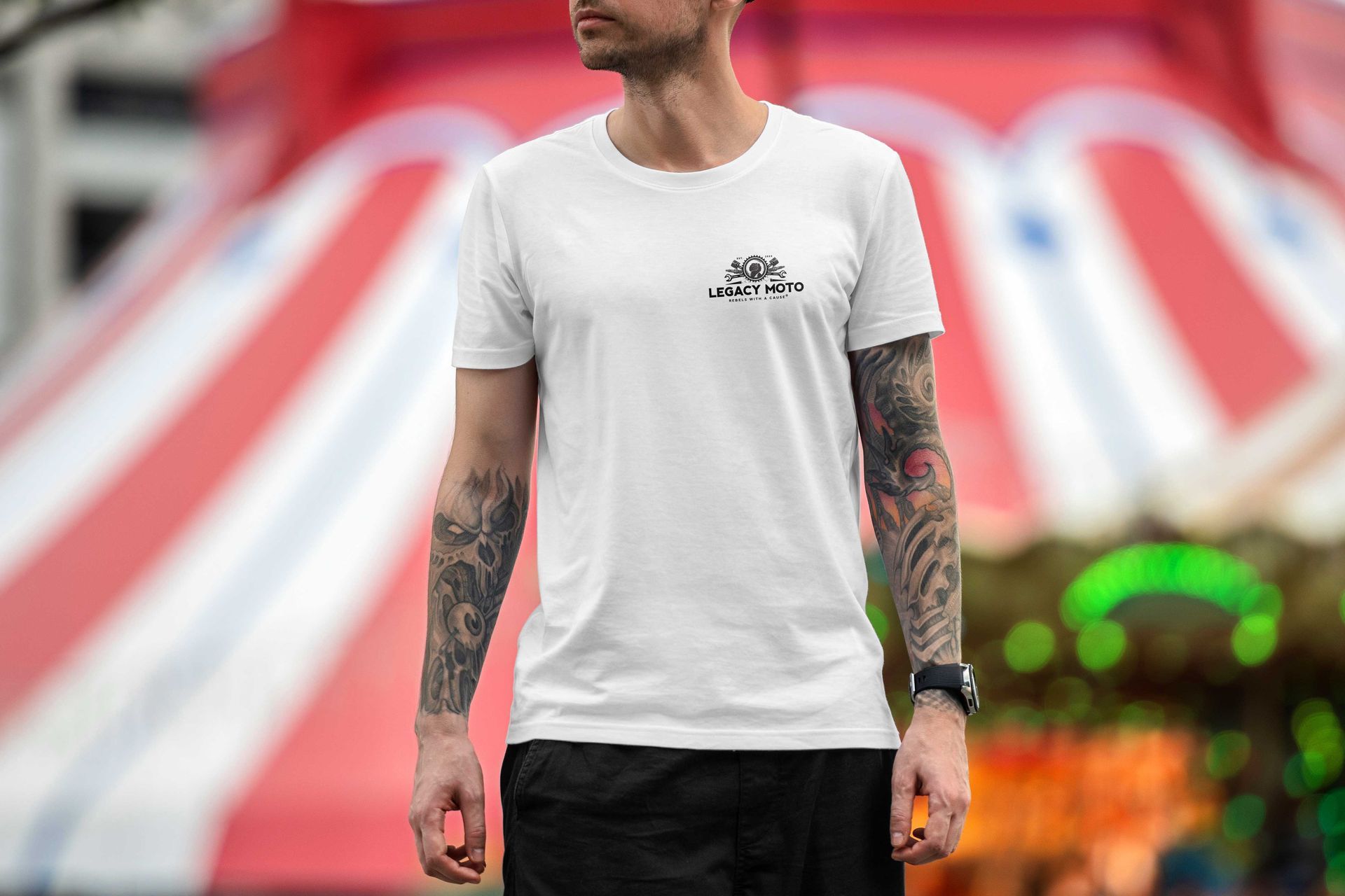 Tattooed person in a white T-shirt standing before a blurred stadium backdrop