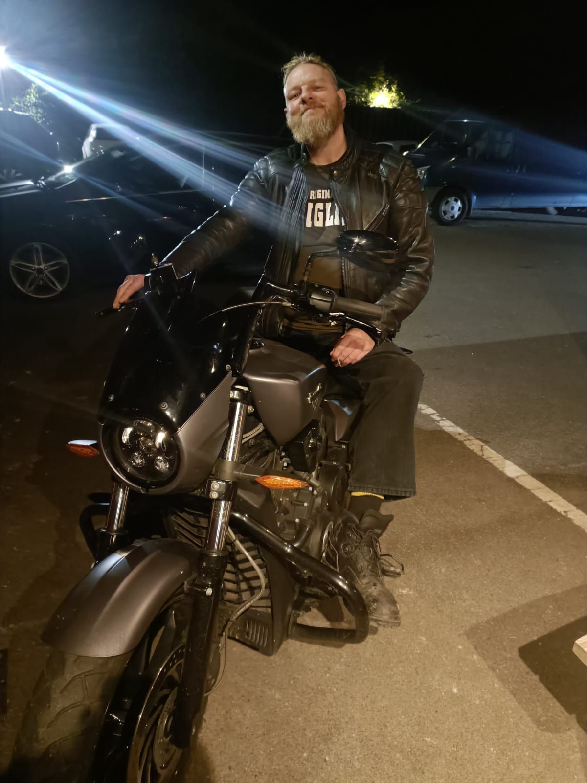 Smiling rider on a black motorcycle at night in a parking lot, wearing a black jacket.