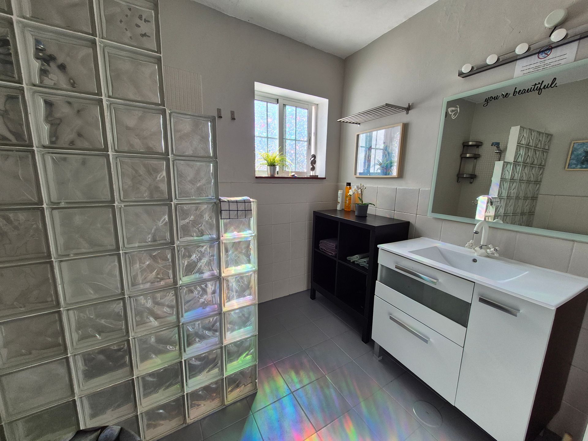 Das Badezimmer mit Waschbecken, Spiegel und Dusche mit Glasbausteinen.