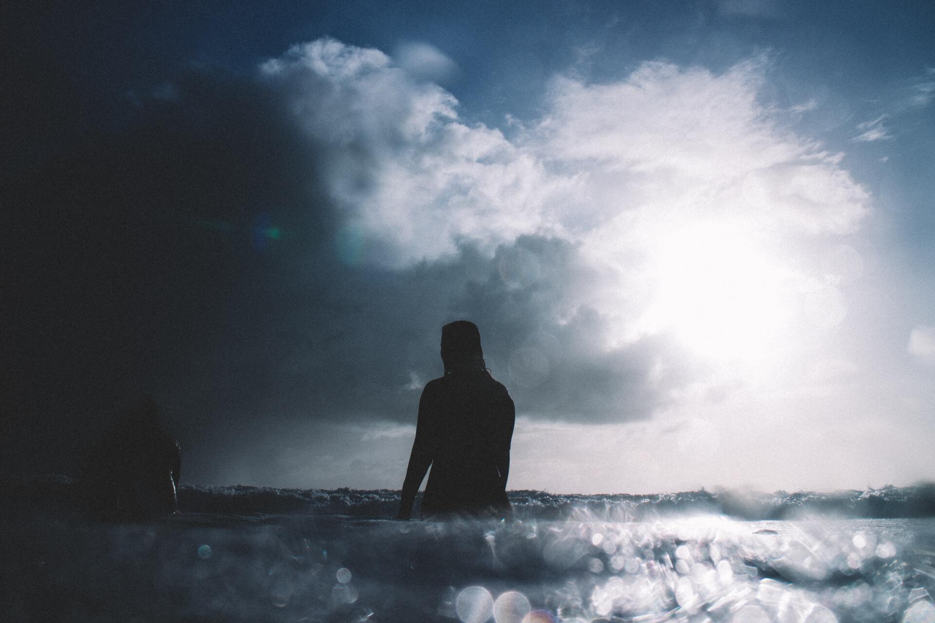 Eine Person sitzt auf einem Surfbrett im Meer, im Hintergrund ist ein bewölkter Himmel zu sehen.