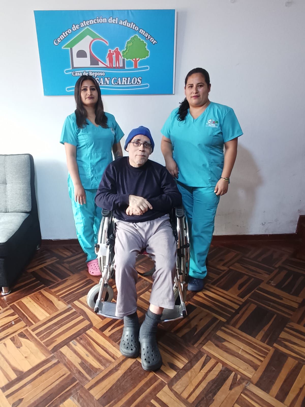 Tres mujeres posando en el interior, una de ellas sentada en una silla de ruedas, con un cartel azul en la pared detrás.