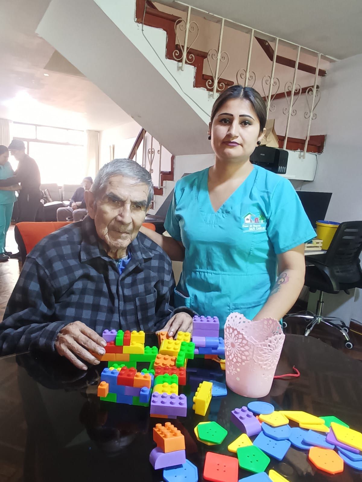 Dos personas sentadas a una mesa con bloques de colores y piezas de rompecabezas en una habitación interior luminosa.