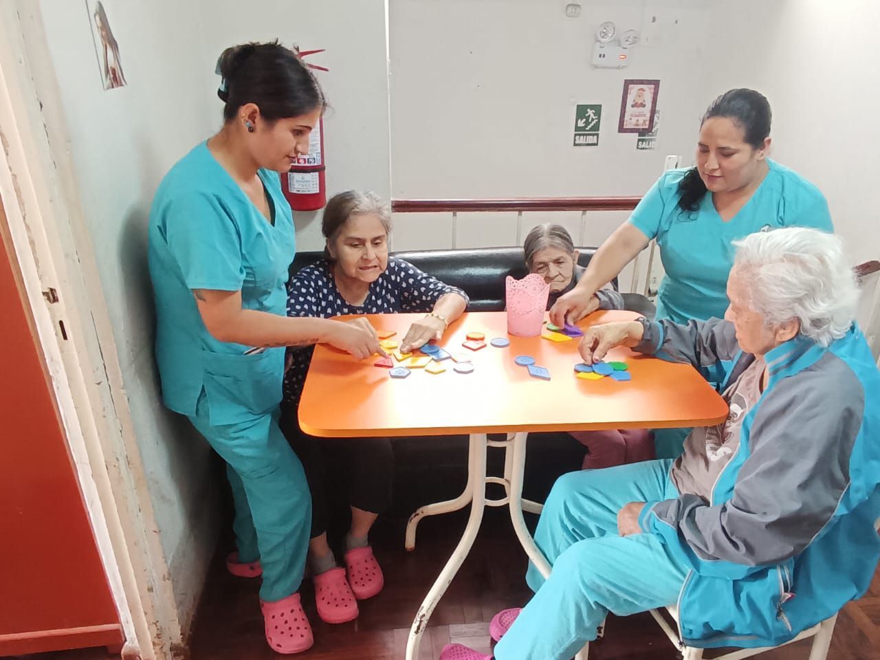 Un cuidador ayuda a tres personas en una mesa naranja en una habitación pequeña, donde un niño y adultos mayores juegan