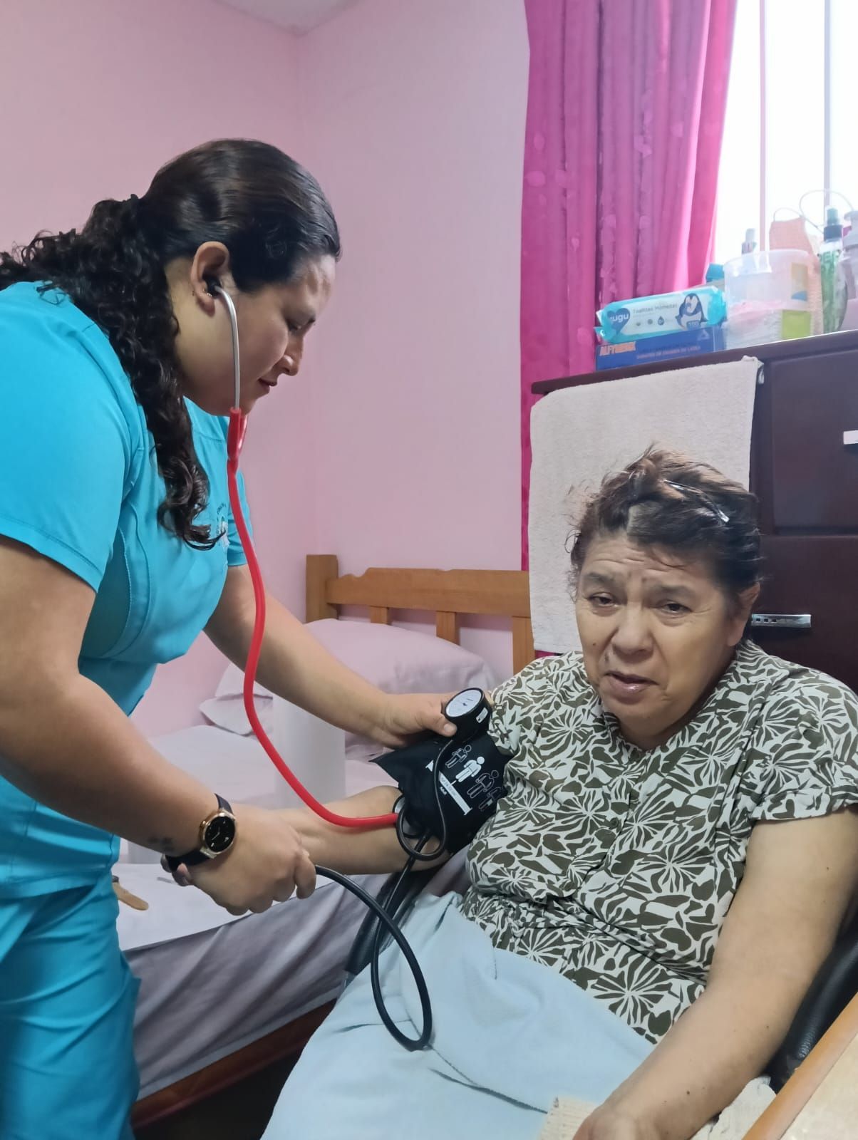 Enfermera tomando la presión arterial a un paciente en una habitación rosa; el paciente parece incómodo.