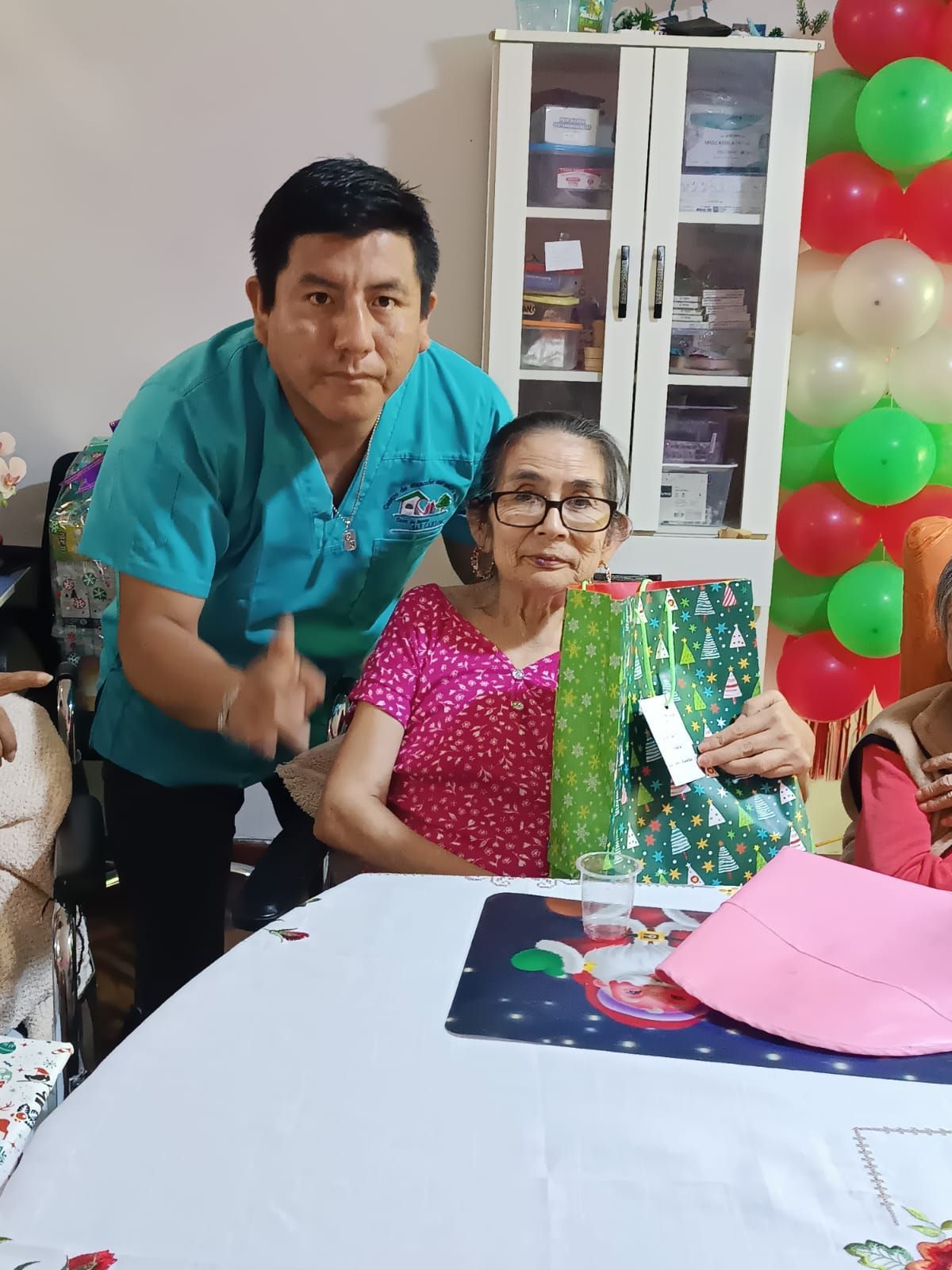 Dos personas sentadas a una mesa con regalos y globos, posando para una foto de celebración.