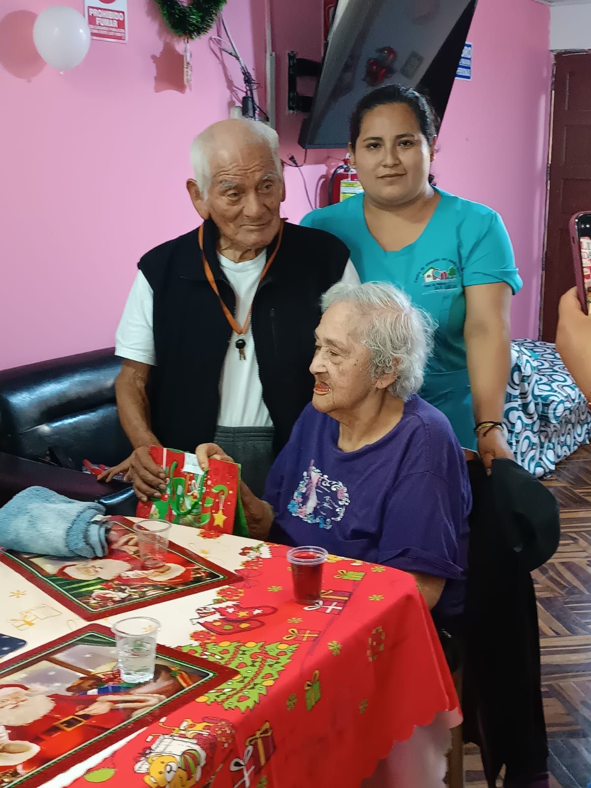 Tres personas celebrando en una mesa con un mantel rojo de cumpleaños y un pastel en una habitación rosa.