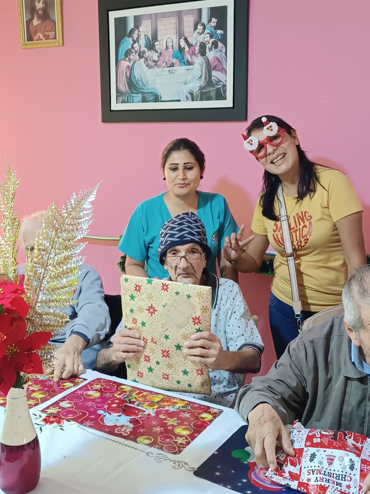 Tres personas sentadas a una mesa abriendo regalos en una sala de estar rosa, sonriendo y posando para una celebración.