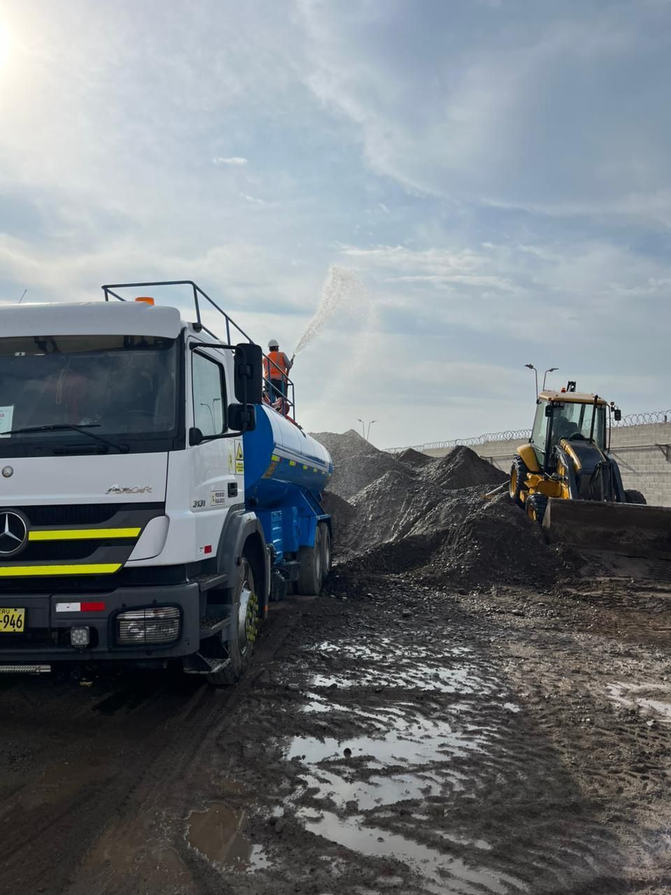 Camiones de construcción en un camino embarrado junto a montones de grava bajo un cielo nublado.