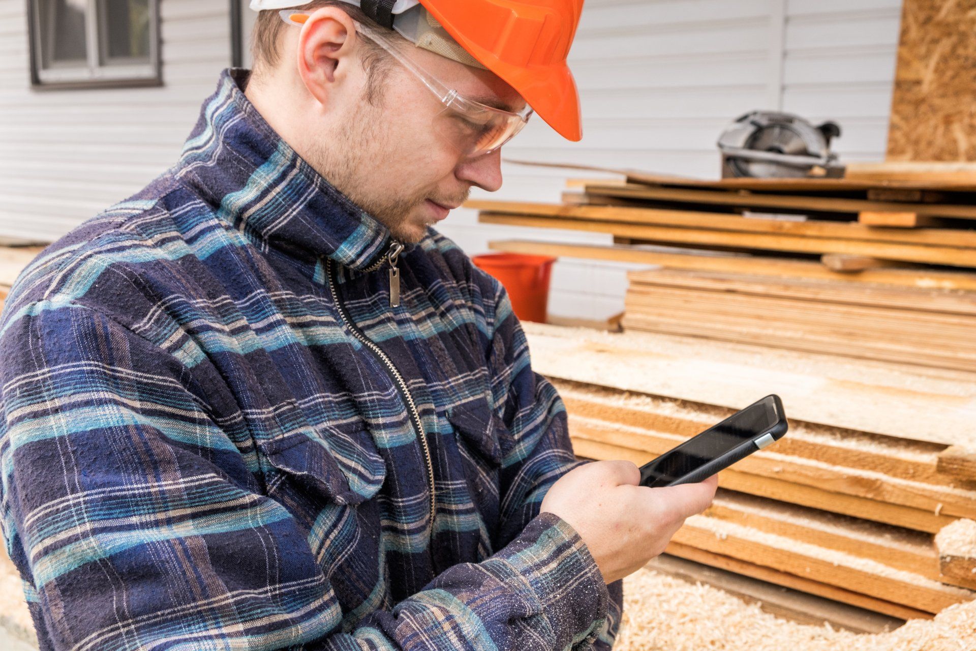 Un ouvrier du bâtiment en chemise à carreaux, casque orange et lunettes de sécurité regarde son téléphone près du bois.