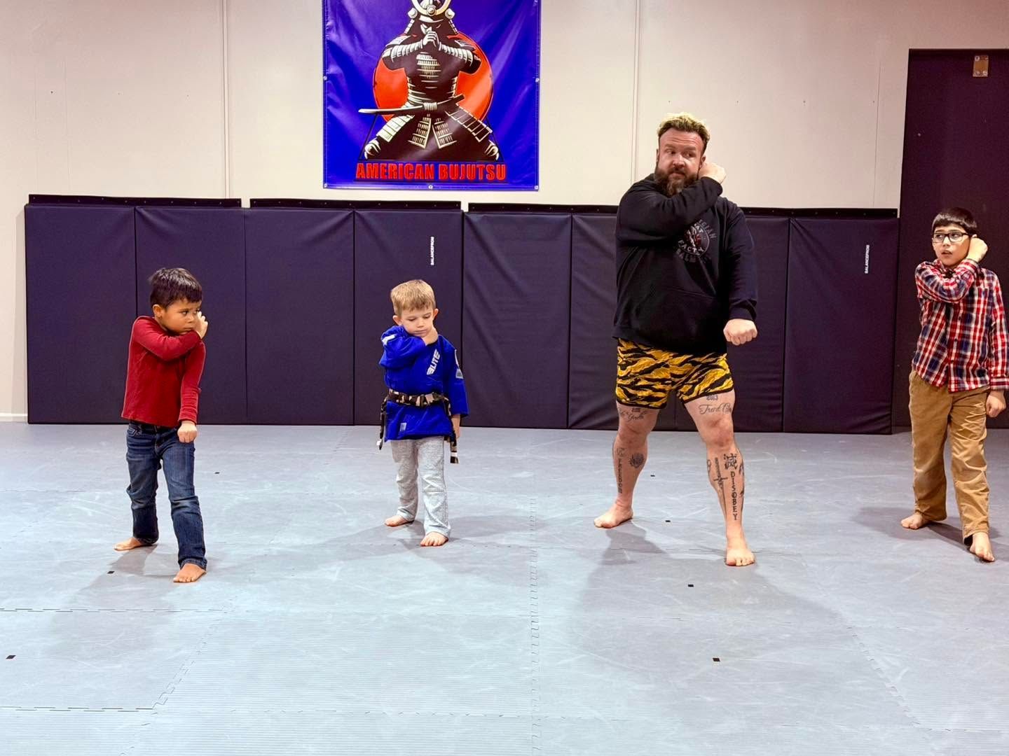 Four people standing barefoot in a martial arts studio, with a coach in black demonstrating a pose.
