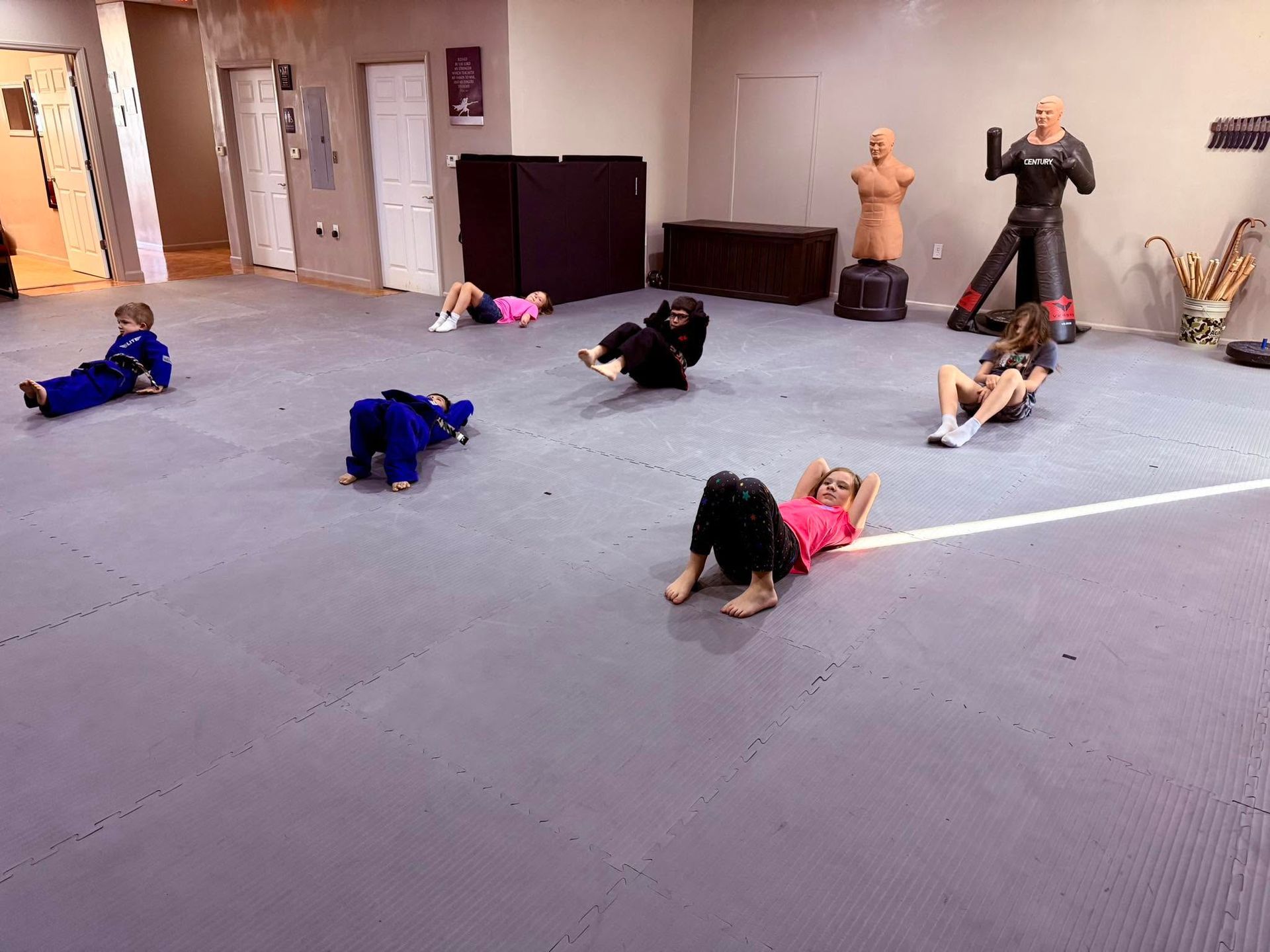 People stretching on mats in a studio, with training dummies and an instructor nearby.